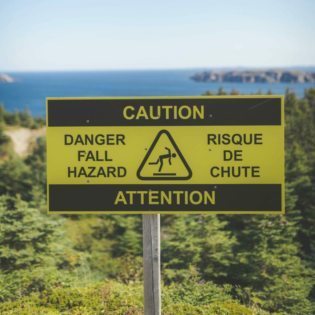 pexels photo 9610455 9610455 Yellow caution sign warns of fall hazard near a scenic ocean view with green foliage.